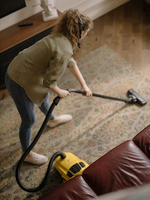 A person using a red vacuum cleaner to clean a beige textured carpet in a living room, with part of wooden furniture visible on the left and natural light illuminating the space. The vacuum cleaner is