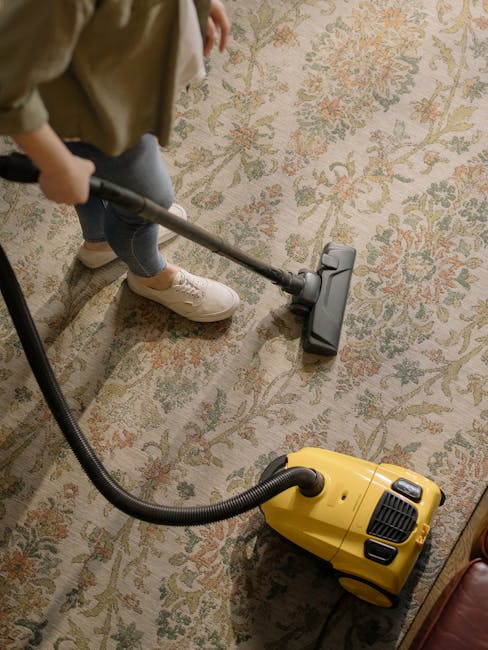 A person operating a yellow vacuum cleaner with a black hose attachment on a traditional patterned, cream-colored carpet in a room with wooden flooring visible at the edges. The individual is wearing