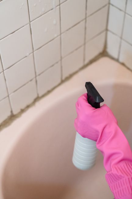 A person with a light pink cropped top and grey sweatpants is engaged in surface cleaning within a bright, well-lit room. They are wiping a wooden table with a white cloth, which appears polished and